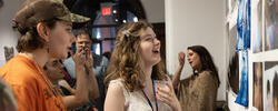 two students look at works on paper mounted on gallery wall as other people move through the gallery