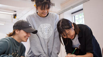 three students smiling together, at a desk while one draws