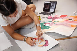 a student sits on a studio floor composing colorful work on paper