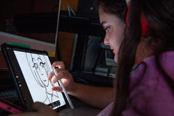 a student draws a cartoon face on a white digital tablet inside a dark room
